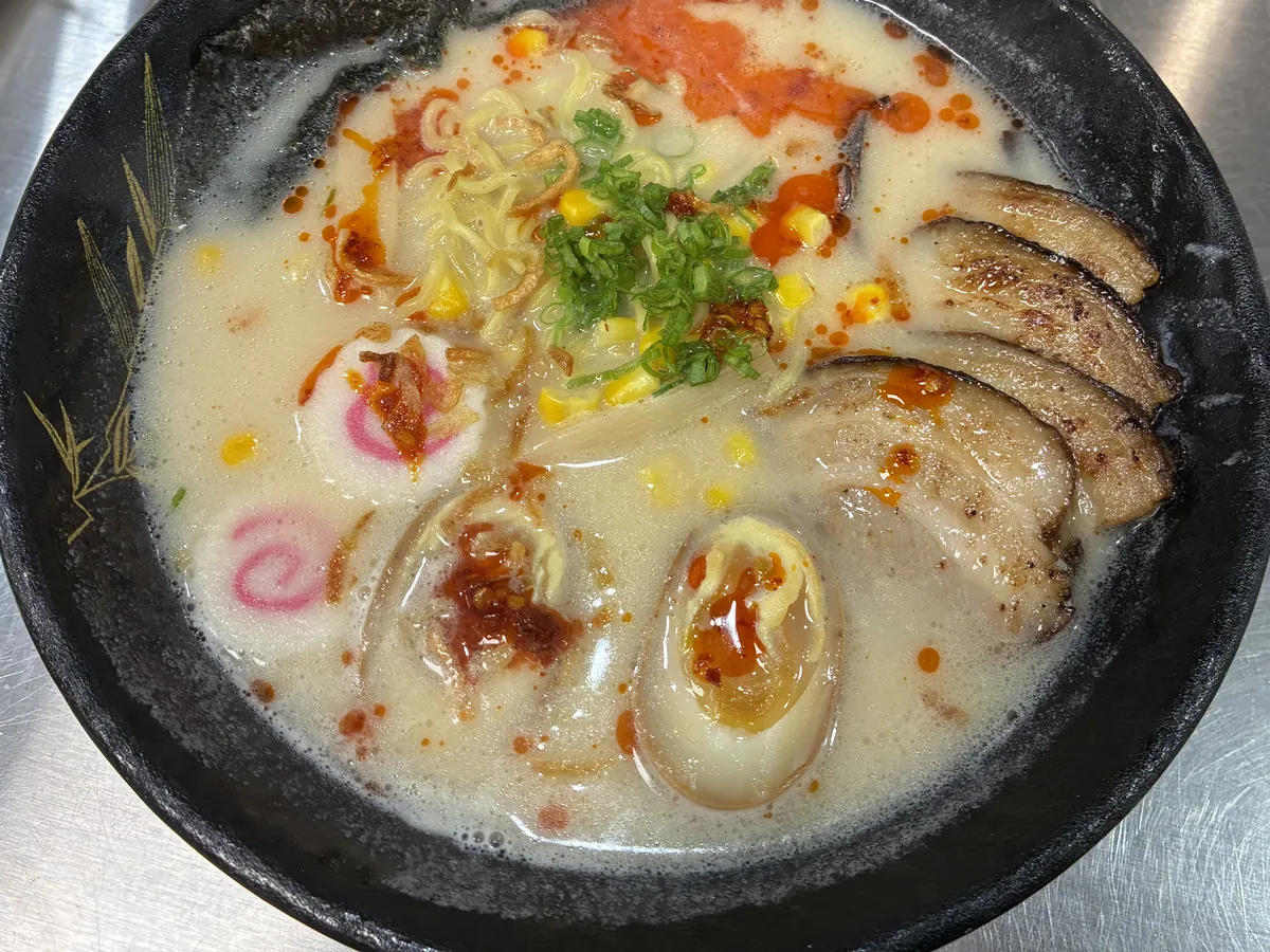 Spicy tonkotsu ramen photographed on the steel pass, chilli oil pooled across the broth over chashu pork and a soft egg
